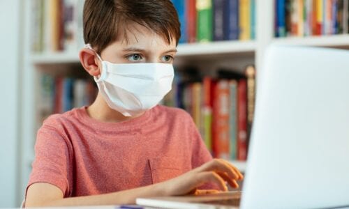 Boy wears a face respiratory face mask while working on schoolwork on his laptop