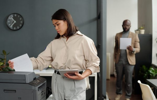 Young serious businesswoman with tablet taking printed paper document from xerox machine while standing against coworker and using tablet